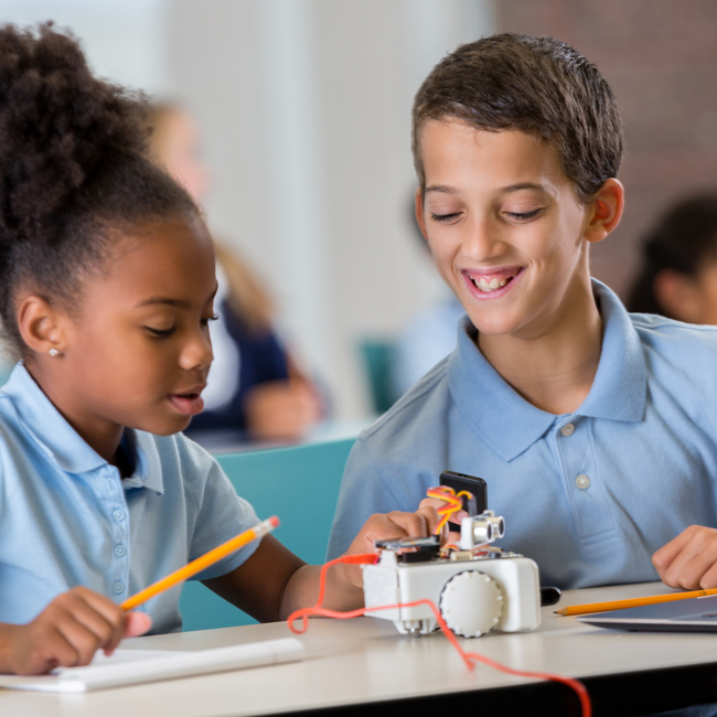 Primary school pupils in a classroom working with a laptop and a working model. One holds a pencil and presses a button on the model while the other watches and smiles.