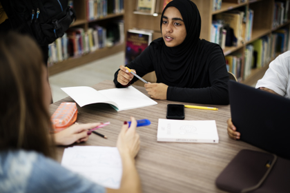 Three secondary school students working at a table in a school library. They are surrounded by books on shelves. On the table they have books and notebooks as well as pens and a pencil case.