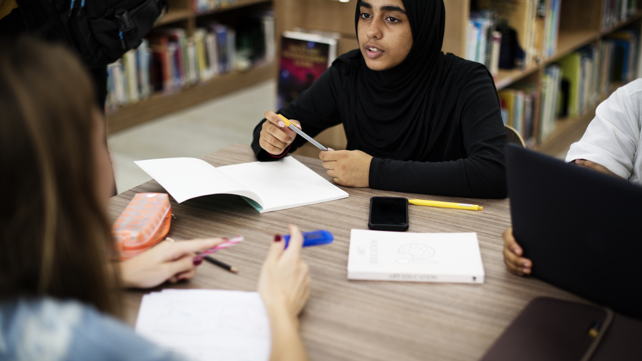 Three secondary school students working at a table in a school library. They are surrounded by books on shelves. On the table they have books and notebooks as well as pens and a pencil case.