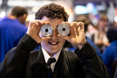 A young person at The Big Bang Fair holds two metal cylindrical pieces in front of their eyes as they smile. 