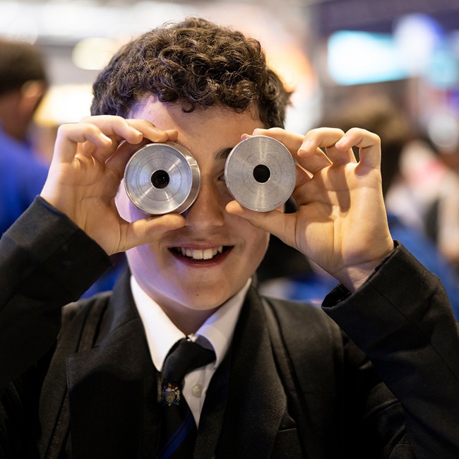 A young person at The Big Bang Fair holds two metal cylindrical pieces in front of their eyes as they smile. 