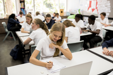 A classroom of secondary school students working at their desks with papers and pencils and laptops. 