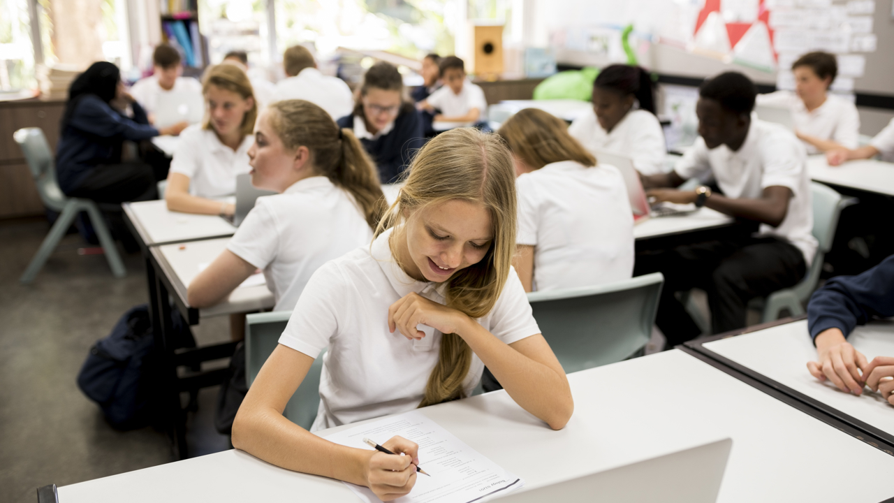 A classroom of secondary school students working at their desks with papers and pencils and laptops.