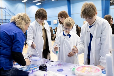 A school science fair setting. Four young people visit a stand and are conducting an experiment supported by a grown up. They all wear protective clothing and goggles. They work with electronic pipettes and liquids.