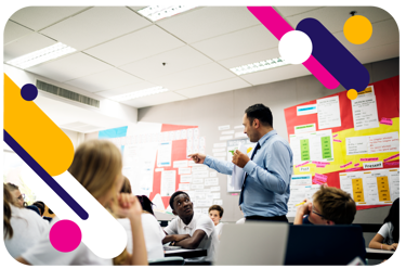 Young people in a classroom. They look up at the teacher, standing and pointing. 