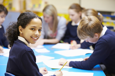 A group of primary school pupils work around a table with notebooks and pencils. One turns and smiles at the camera. A teacher is sitting with them visible in the background.