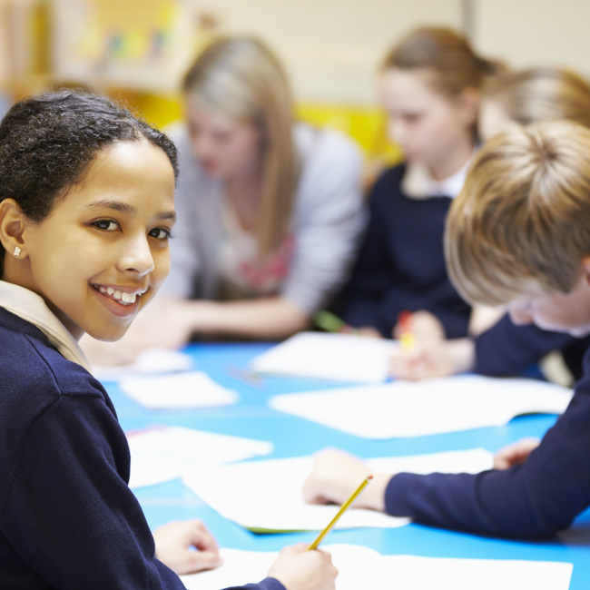 A group of primary school pupils work around a table with notebooks and pencils. One turns and smiles at the camera. A teacher is sitting with them visible in the background.