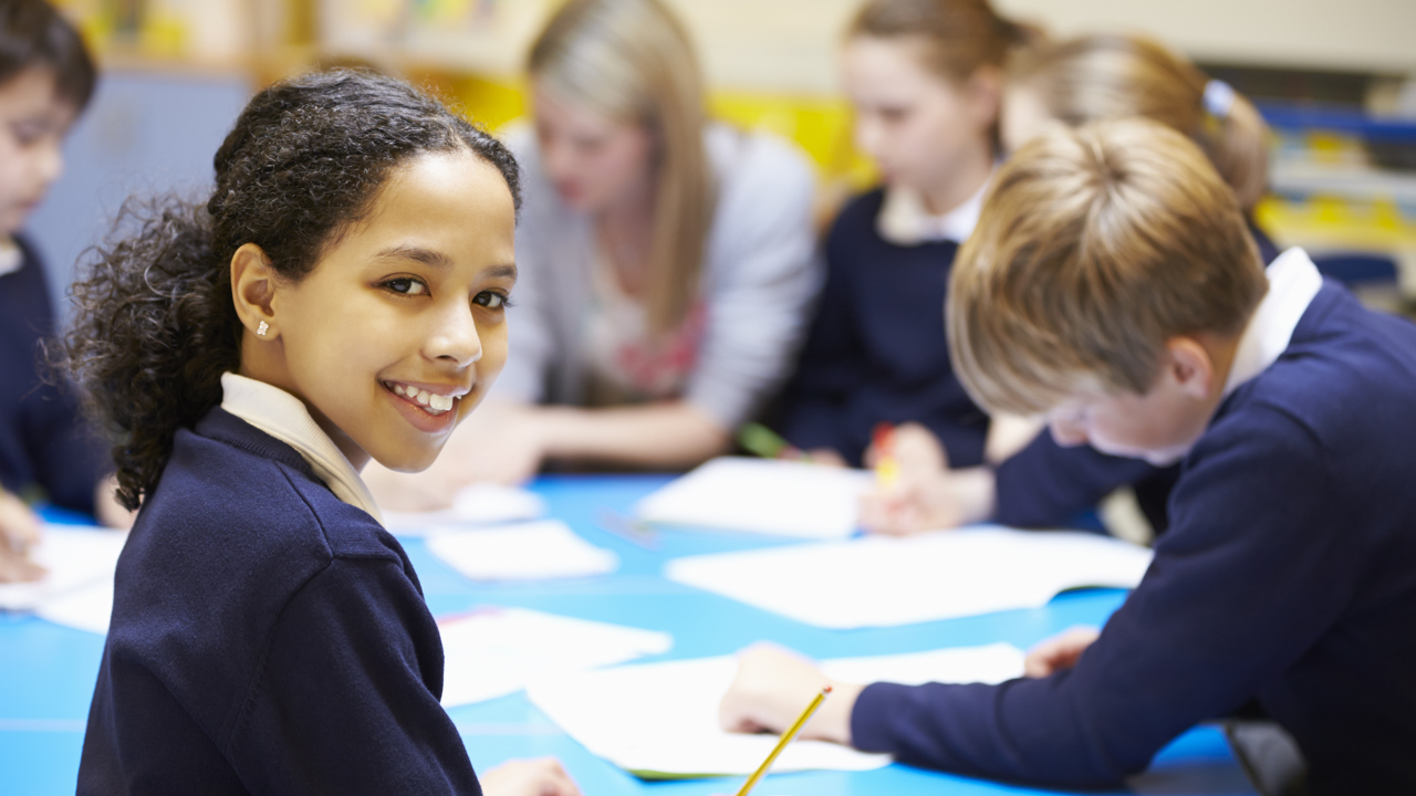 A group of primary school pupils work around a table with notebooks and pencils. One turns and smiles at the camera. A teacher is sitting with them visible in the background.