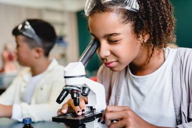 Young person looking down a microscope in a school science laboratory. A second person is seen in the background with goggles on.