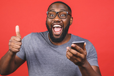 A young adult holds a phone and the other hand has a thumbs up. They smile. 