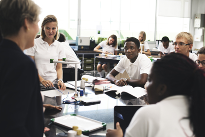 A science classroom in a secondary school. A teacher talks to a group of students who are sitting and standing around a work bench, listening. On the work bench is scientific equipment and workbooks. 