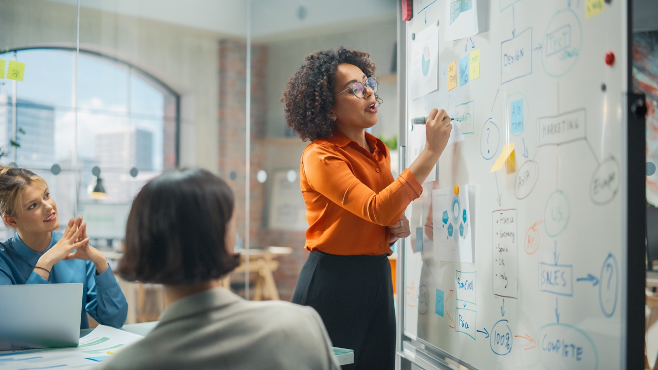 Three people at work in a meeting. One is standing and writing at a whiteboard while the other two look on.