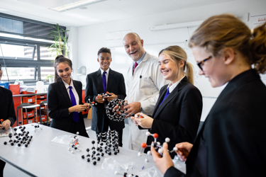 A teacher with a selection of pupils in a secondary school science lesson. They stand around a table which has lots of models on it. Some are holding and interacting with the model structures. 