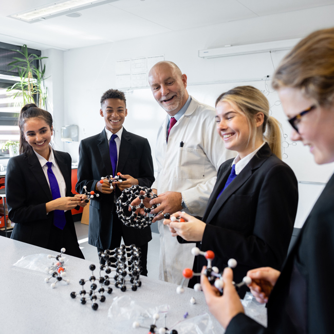 A teacher with a selection of pupils in a secondary school science lesson. They stand around a table which has lots of models on it. Some are holding and interacting with the model structures. 