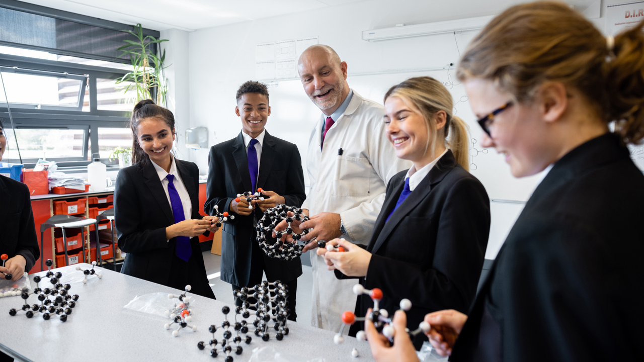 A teacher with a selection of pupils in a secondary school science lesson. They stand around a table which has lots of models on it. Some are holding and interacting with the model structures. 