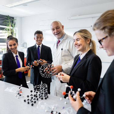 A teacher with a selection of pupils in a secondary school science lesson. They stand around a table which has lots of models on it. Some are holding and interacting with the model structures.