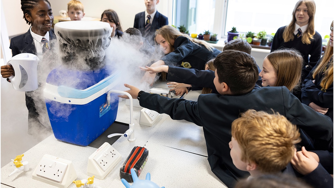 A school science lab. Many students surround a work bench as a cool box exudes dry ice. The students reach to touch it. They smile as they watch on. 