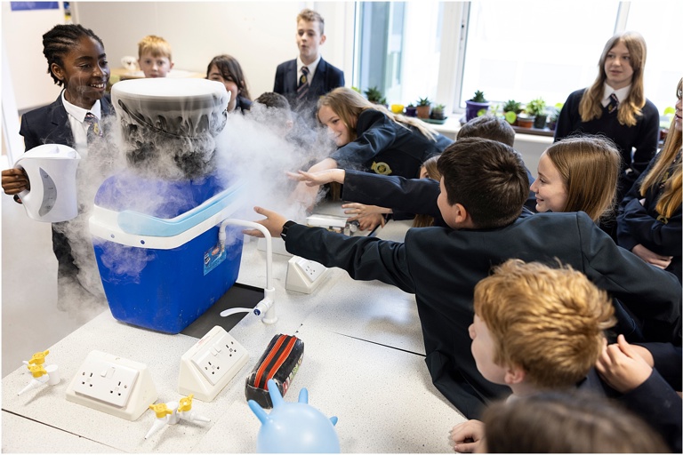 A school science lab. Many students surround a work bench as a cool box exudes dry ice. The students reach to touch it. They smile as they watch on.