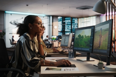 A worker sits at their desk in front of two monitors. They are staring at a screen and holding a mouse.