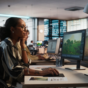 A worker sits at their desk in front of two monitors. They are staring at a screen and holding a mouse.