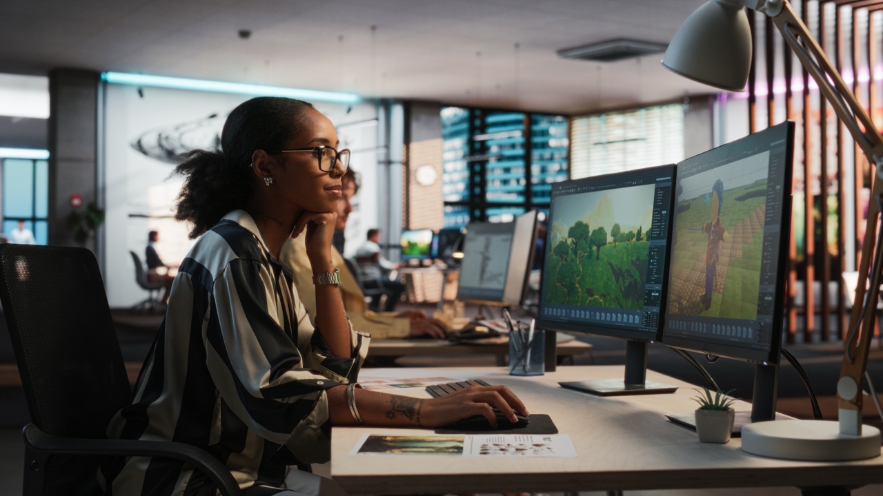 A worker sits at their desk in front of two monitors. They are staring at a screen and holding a mouse.