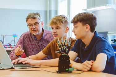 Two young people and their teacher sit in a school classroom looking at a computer as they study how to code a model robot, also on their desk