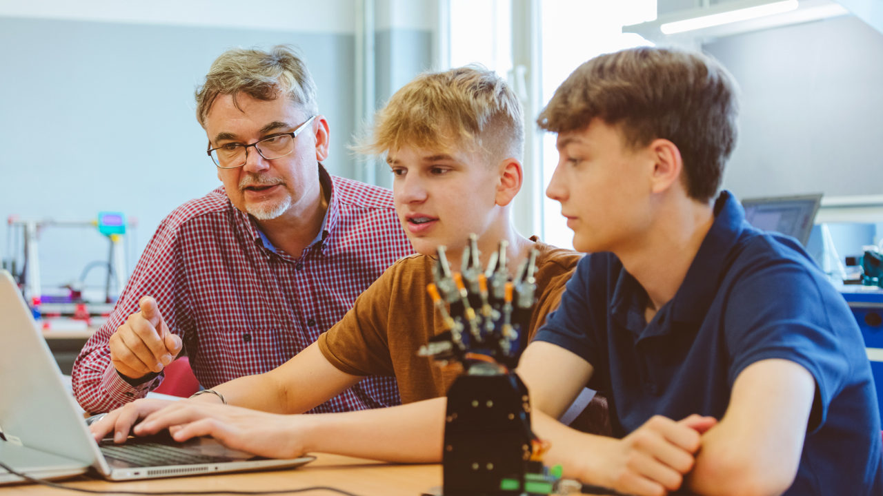 Two young people and their teacher sit in a school classroom looking at a computer as they study how to code a model robot, also on their desk