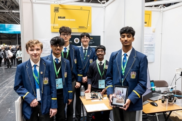 The six winners of The Big Bang Competition UK Young Technologists of the Year stand in an exhibition area displaying a model of their product along with sketches and working drawings to show how their idea developed. They all face the camera