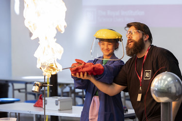 A young person and a teacher stand together conducting a scientific experiment with fire. The student has protective gloves, goggles and overall. They are holding something in the flame as the grown up supports them.