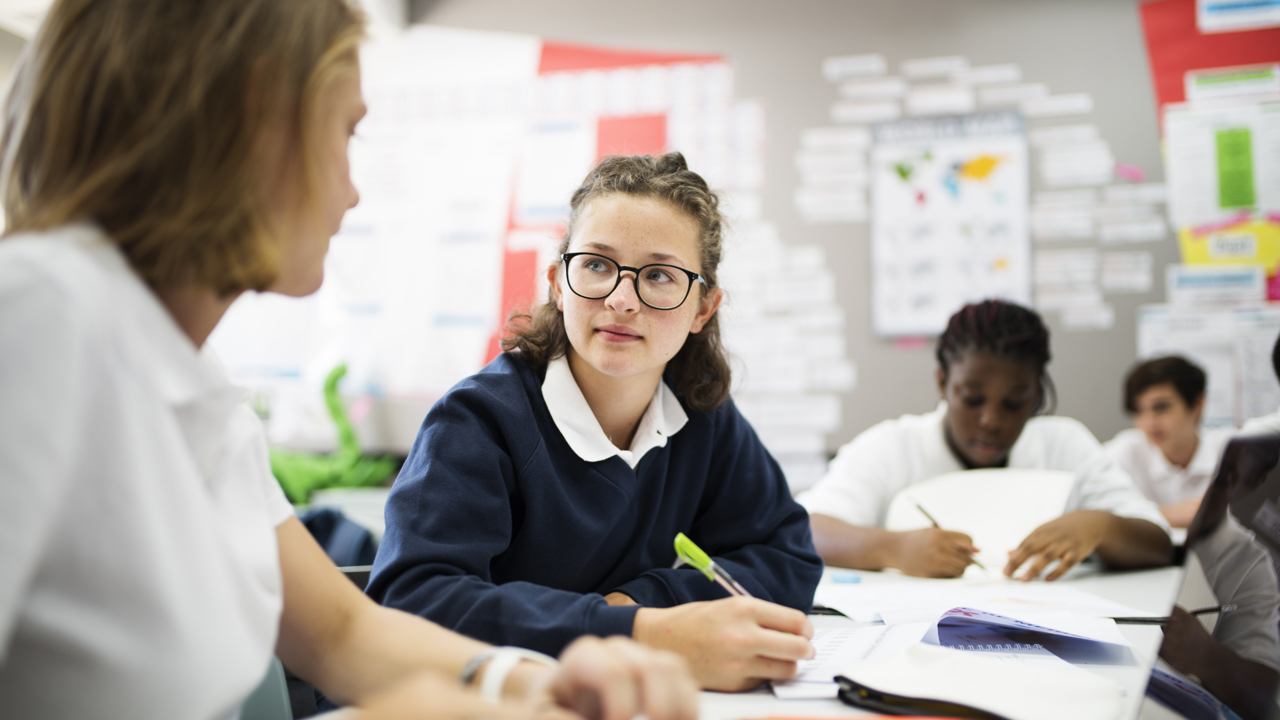 A group of young people work in a classroom. They have pens and notebooks.