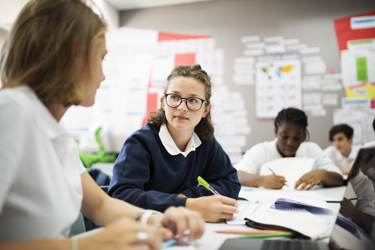A group of young people work in a classroom. They have pens and notebooks.