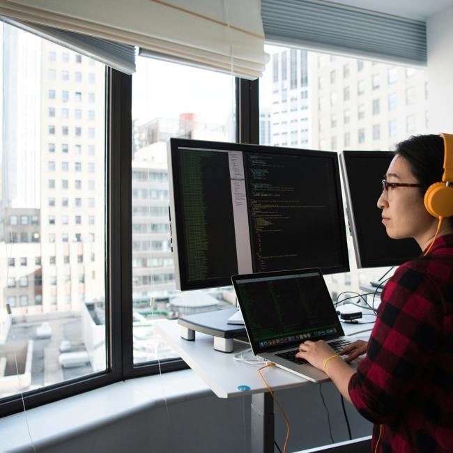 An adult sits at a desk with headphones on. They are typing on their laptop and looking at three screens. They are high up and outside their window you can see other buildings 