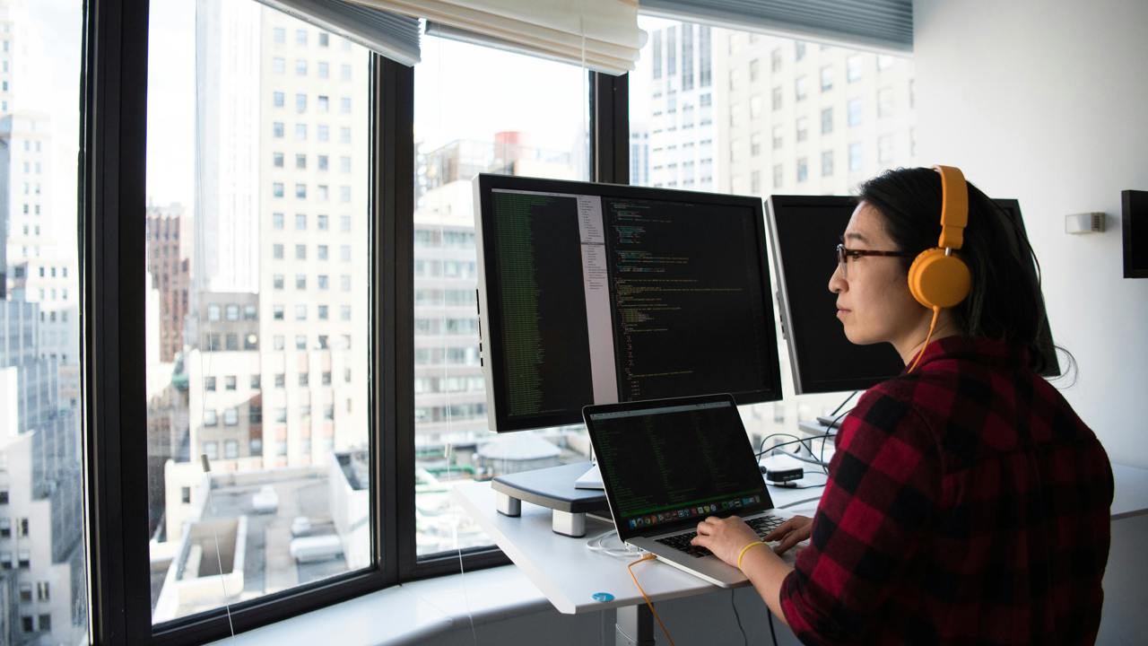 An adult sits at a desk with headphones on. They are typing on their laptop and looking at three screens. They are high up and outside their window you can see other buildings 