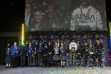A group shot of The Big Bang Competition winners all together on a stage holding their awards. A confetti canon has released ticker tape which is still floating in the air, and some has rested on the stage.