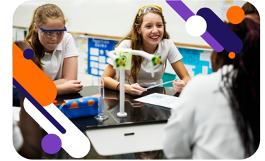 A group of girls in a school science lab smiling as they work at the bench. They are wearing goggles. 